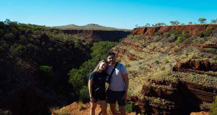 Lachendes Paar posiert am Rand einer dramatischen roten Felsenschlucht unter einem klaren blauen Himmel in Westaustralien.