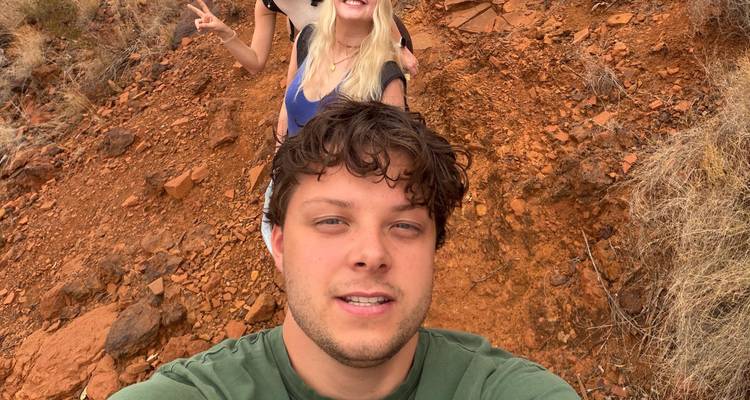 Close selfie of a traveller on a red dirt trail with a friend flashing a peace sign behind.