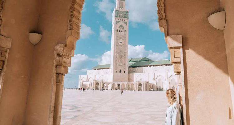 Eine weibliche Reisende lehnt sich an einen Bogen und rahmt den Innenhof der Hassan-II-Moschee unter blauem Himmel ein.