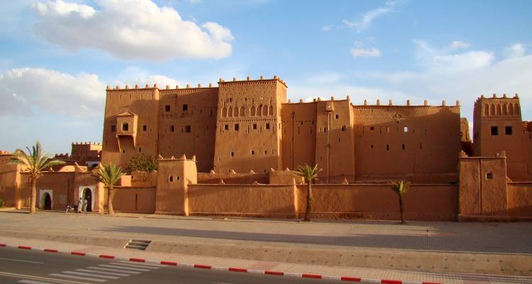 Immense kasbah de couleur ocre avec des murs crénelés et de petits palmiers sur un ciel bleu.