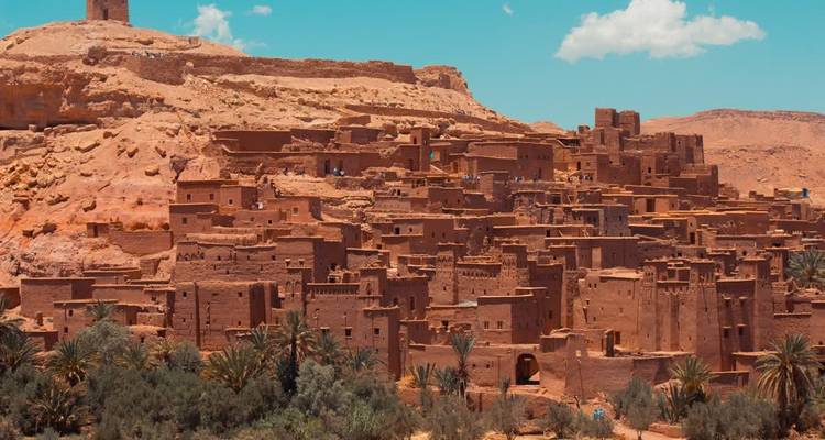 Les structures de kasbah en terre d'Aït Benhaddou s'étagent sur une colline au milieu des palmiers sous un ciel bleu.