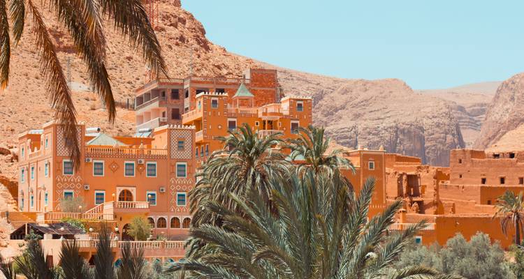 Un village fortifié de couleur ocre aux architectures marocaines complexes encadré par de grands palmiers et des montagnes désertiques arides.