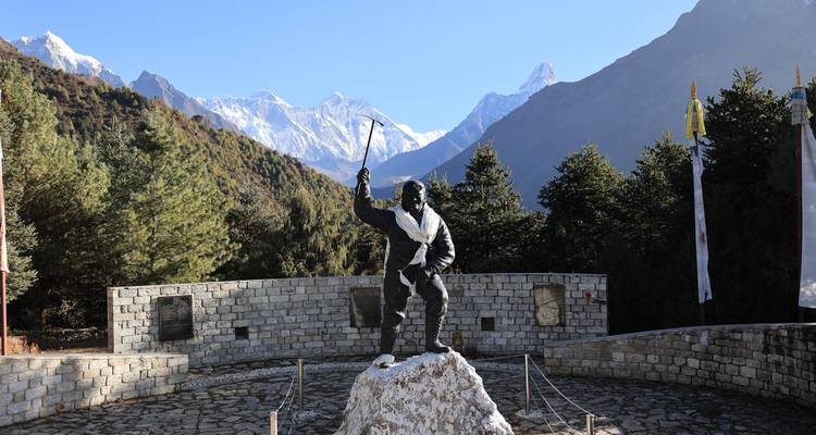 Bronzen standbeeld van Tenzing Norgay met een ijsbijl in de hand, met op de achtergrond beboste Himalayatoppen en de verre Everest-bergketen.