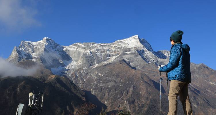 Solo wandelaar met wandelstokken kijkt omhoog naar een robuuste sneeuwbedeekte Himalaya-kam tegen een wolkenloze hemel.