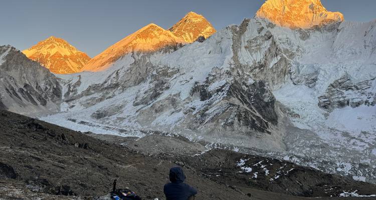 Een wandelaar rust uit op een rotsachtige helling en kijkt hoe het gouden zonnlicht de Everest, Nuptse en naburige bergtoppen boven een gletsjer schildert.