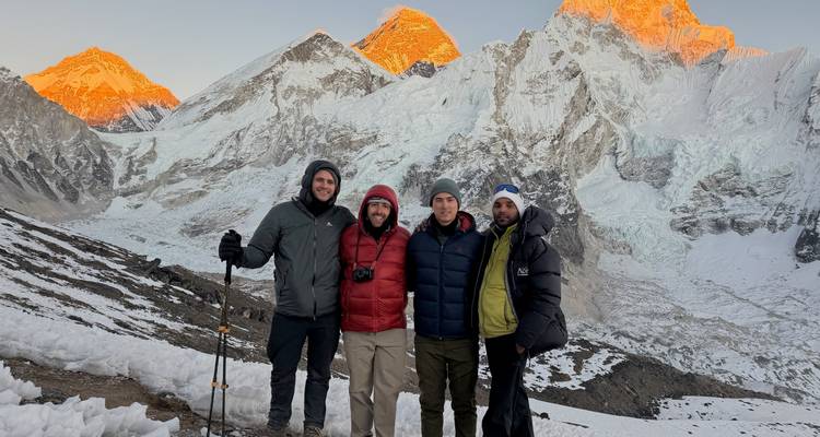 Vier lachende trekkingtoeristen poseren op een sneeuwrichel met glooiende oranje Everest-toppen achter hen in het schemerlicht.