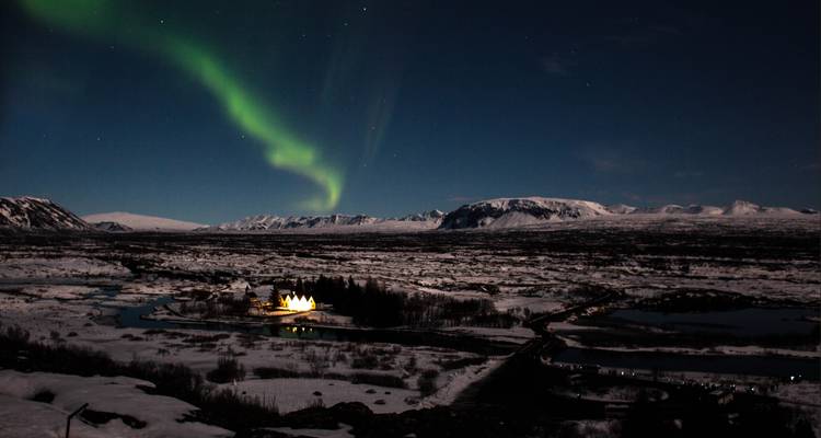 Auroras boreales sobre un paisaje nevado con un río, montañas y una casa iluminada.
