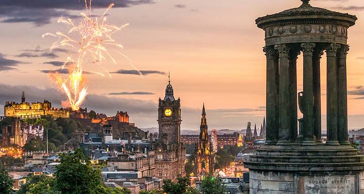 Fireworks explode over Edinburgh Castle with the Dugald Stewart Monument and city skyline glowing at sunset
