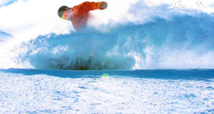 Photographie d'action d'un snowboardeur traçant sa courbe dans la poudreuse fraîche, projetant une gerbe de neige sur un ciel bleu éclatant.