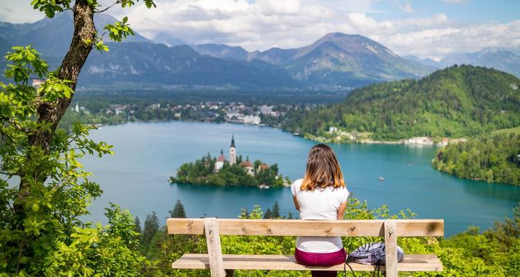 Mujer sentada en un banco de madera con vistas al lago turquesa de Bled, su iglesia en la isla y las montañas que lo rodean.