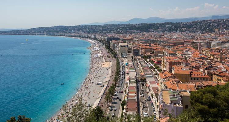 Sweeping aerial of Nice’s curving pebbled beach and azure Mediterranean backed by the city and Alps.