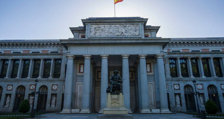 Front façade of Madrid’s Prado Museum with statue and Spanish flag under bright backlight.