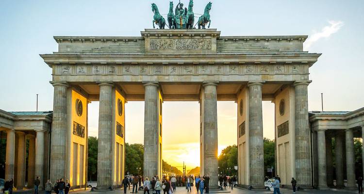 La Puerta de Brandeburgo iluminada por la luz dorada de un atardecer cálido mientras multitudes de visitantes pasan bajo sus columnas neoclásicas.