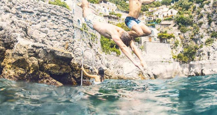 Dos jóvenes se lanzan desde una orilla rocosa hacia las aguas azules y cristalinas del Mediterráneo mientras una mujer espera en una escalera debajo de acantilados junto al mar y casas en terrazas.