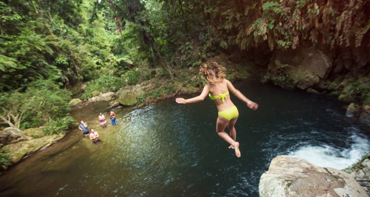 Una mujer en bikini amarillo salta desde una roca en la jungla hacia una piscina cristalina mientras sus amigos la miran desde abajo.