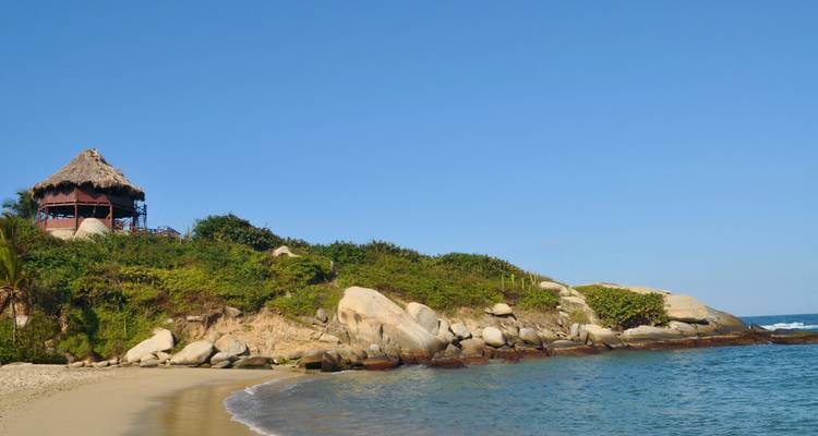 Una pequeña cala de arena con agua turquesa, costa rocosa y una cabaña de techo de paja sentada en una colina verde bajo un cielo azul despejado.