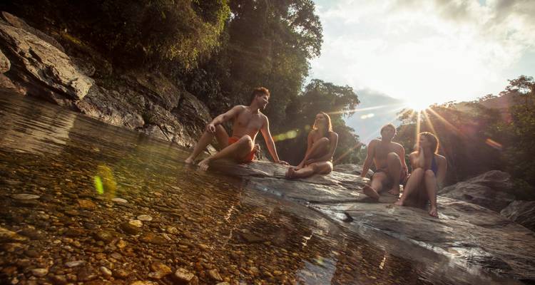 Cinco adultos jóvenes en trajes de baño se relajan en rocas cálidas al lado de un río cristalino mientras el sol se asoma por el dosel del bosque.