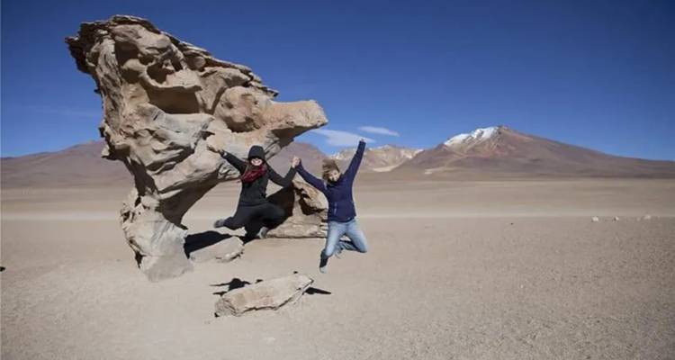 Dos personas saltando cerca de una formación rocosa de forma única en un paisaje desértico.