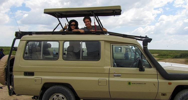 Dos personas de pie en un vehículo safari con techo abierto en una zona silvestre.