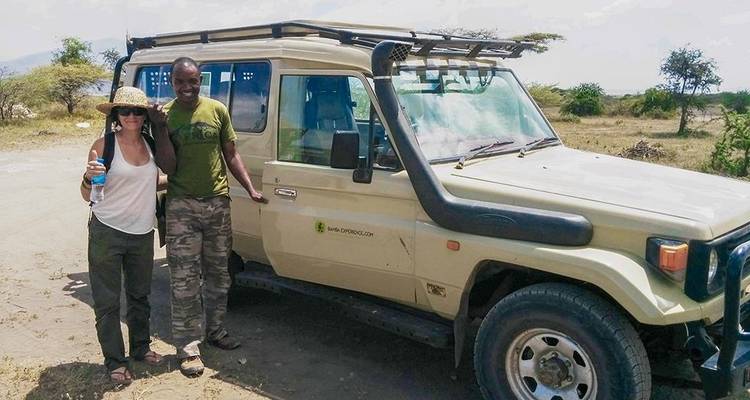 Una mujer y un guía posando frente a un vehículo de safari en una sabana.