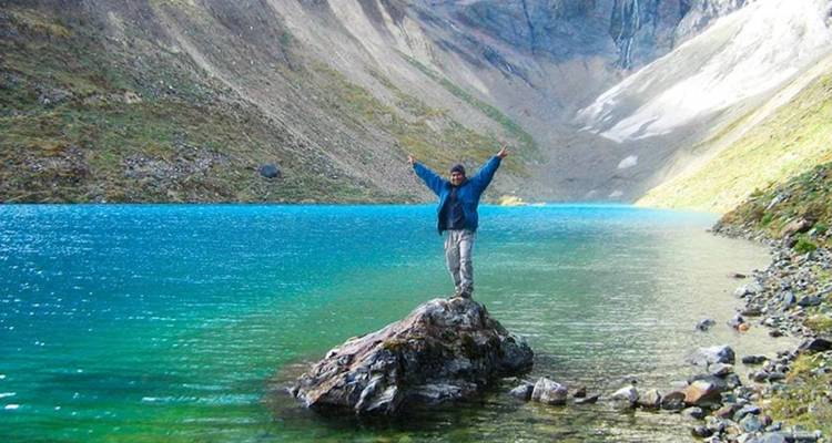 Persona de pie sobre una roca frente a un lago azul rodeado de montañas.