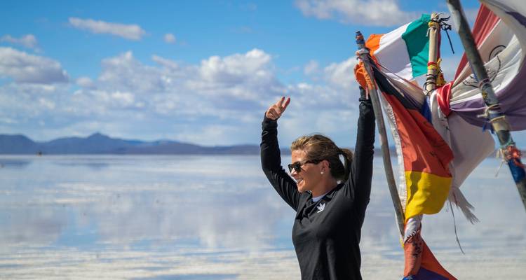 Una mujer de pie con banderas en el Salar de Uyuni con un cielo despejado y agua como un espejo.