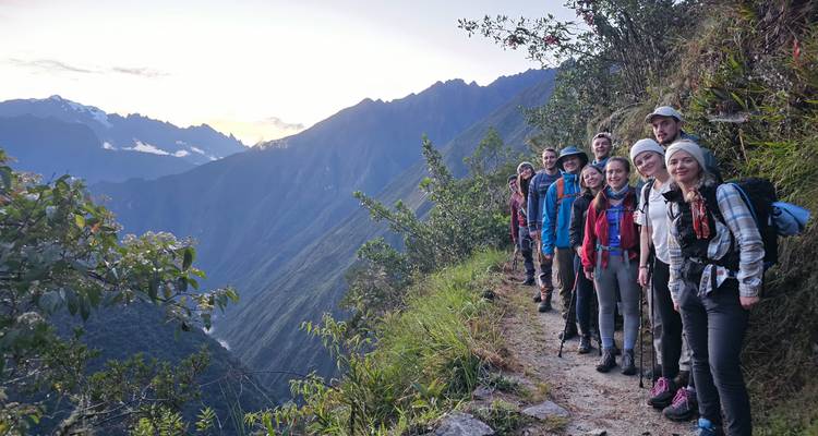 Una línea de excursionistas se para en un estrecho sendero de montaña que da vista a un profundo valle verde mientras el sol sale sobre los picos distantes.