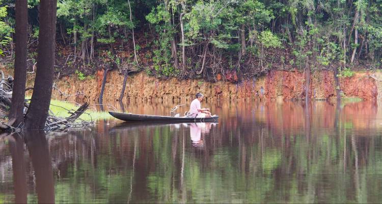 Ein einzelner Paddler in einem kleinen Kanu treibt auf einem spiegelglatten Fluss, der von Dschungel gesäumt wird.