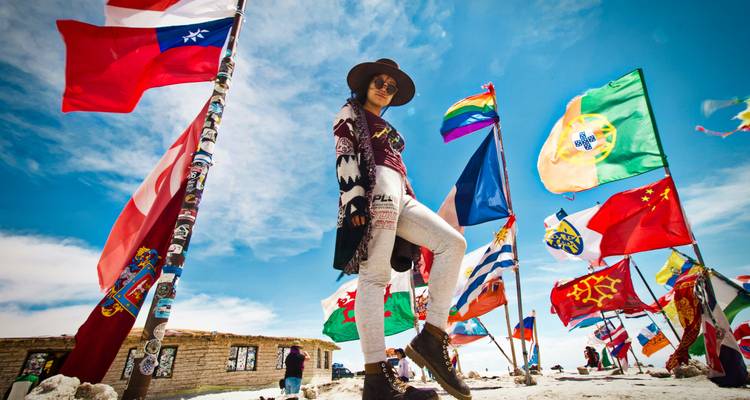 Voyageur posant parmi les drapeaux colorés du monde sur la surface brillante d'une vaste étendue de sel sous un ciel dégagé.