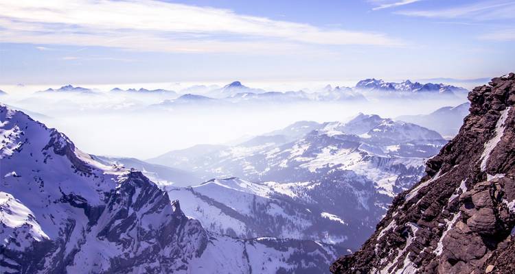 Vue à haute altitude traversant des couches de montagnes alpines enneigées et brumeuses sous un ciel aux teintes pastel.