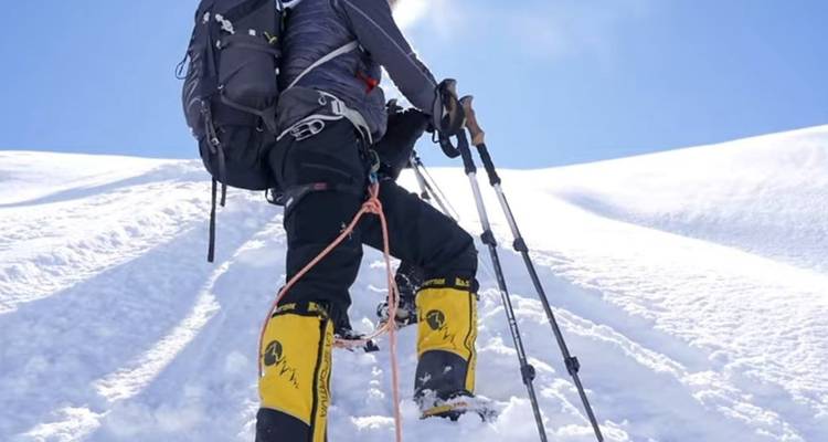 Persoon die besneeuwde berg beklimt met wandeluitrusting onder blauwe hemel.