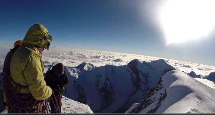 Des personnes en équipement d'escalade debout sur un sommet enneigé avec le soleil qui brille intensément.