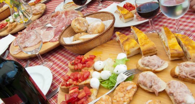 Abundante tabla de antipasti italiana extendida sobre un mantel a cuadros con copas de vino y manos preparando comida.