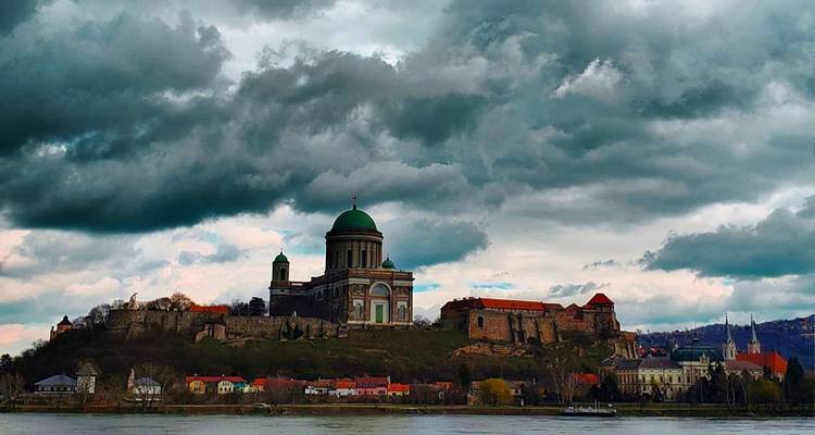Gran basílica en una colina sobre un río bajo nubes de tormenta amenazantes, reflejándose en el agua.