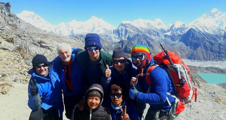 Grupo de excursionistas posando en un terreno rocoso con montañas cubiertas de nieve al fondo.