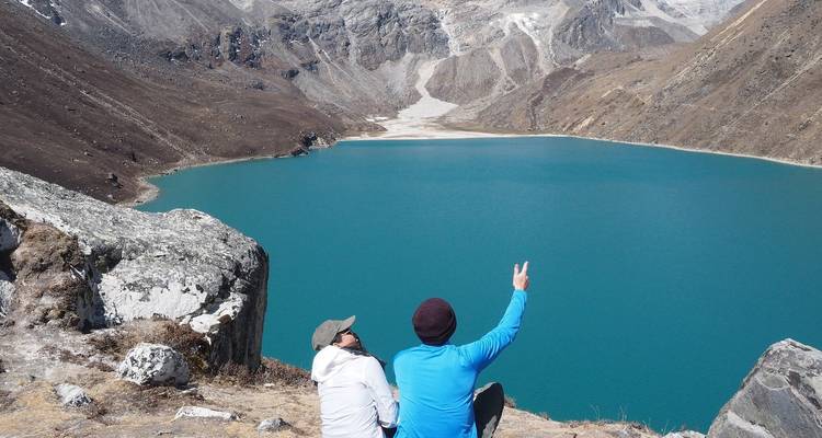 Dos personas sentadas cerca de un lago turquesa vibrante en una zona montañosa.