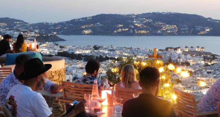 Reizigers genieten van zonsondergang cocktails in een hogeropgelegen bar met uitzicht op de glinsterende lichtjes en baai van Mykonos