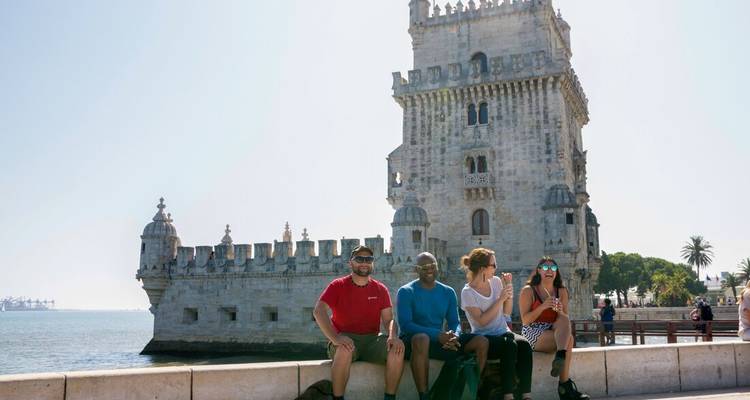 Travellers sit on a riverside wall with Lisbon’s historic Belém Tower rising behind them on a bright day.