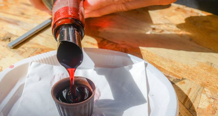 Red ginjinha liqueur pours from a spout into a small chocolate cup on a sunlit counter.