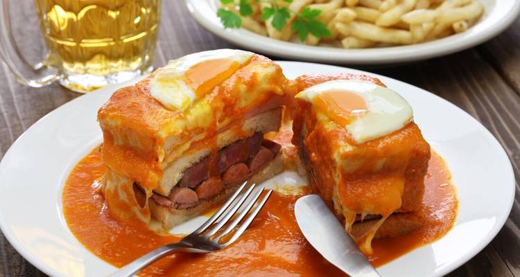 Traditional Porto francesinha sandwich drenched in rich sauce sits cut open on a white plate beside beer.