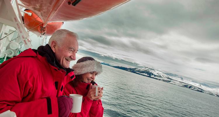 Viajeros ancianos en abrigos rojos beben bebidas calientes en la cubierta de un barco con vista a aguas árticas y picos nevados.