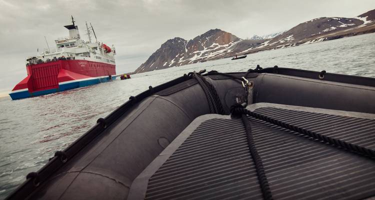 Desde un zodíaco, se ve un barco de expedición rojo cerca de montañas nevadas bajo cielos grises.