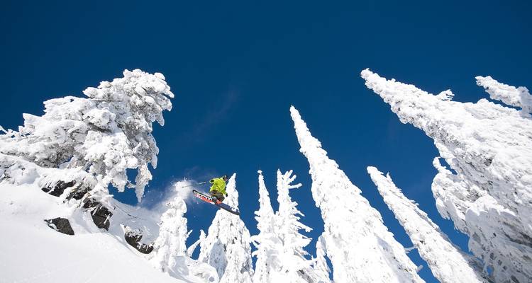 Persona realizando un salto de esquí desde un acantilado nevado bajo un cielo azul.