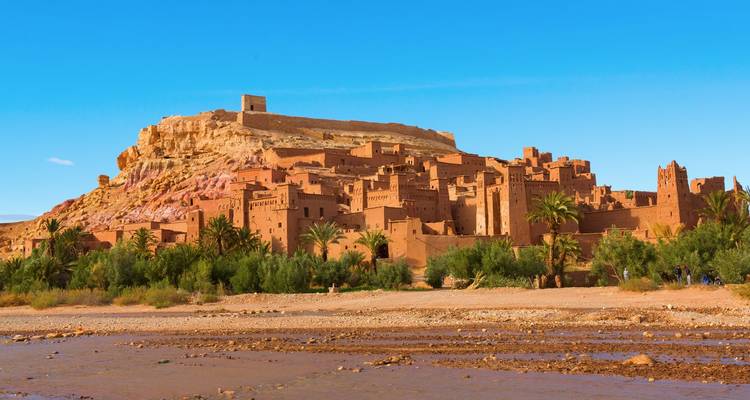 El pueblo de barro fortificado de Ait Benhaddou se alza en lo alto de una colina con un río poco profundo y vegetación en primer plano.