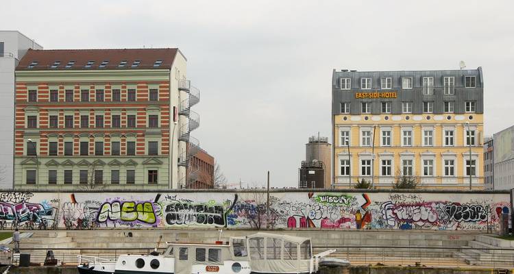 Graffiti-bedekte overblijfsel van de Berlijnse Muur voert langs een waterfront tussen kleurrijke historische gebouwen.