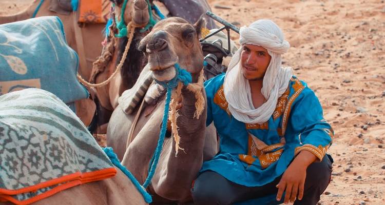 Hombre con ropa tradicional sentado junto a camellos en el desierto.