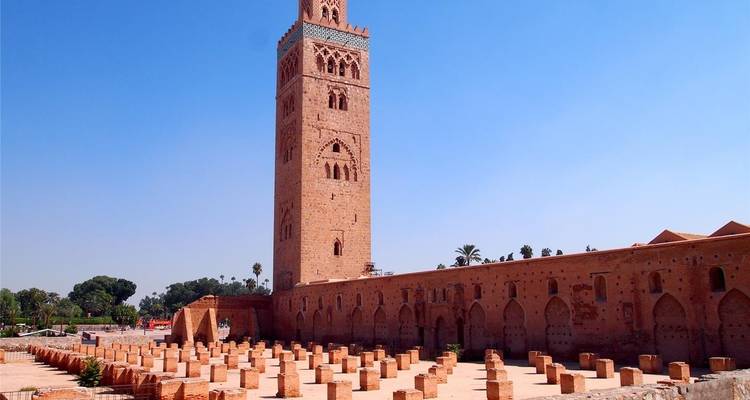 Grande tour de minaret entourée de ruines anciennes, avec des palmiers à proximité.