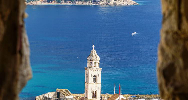 Torre campanario de piedra de una iglesia costera enmarcada por murallas de la ciudad con el vivido mar Adriático azul más allá