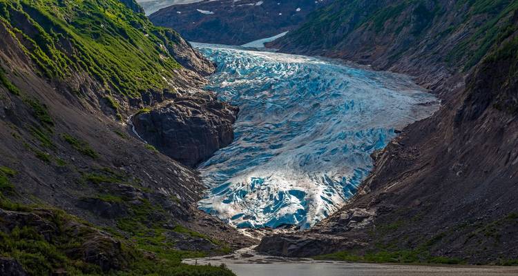 Valle verde y empinado que termina en un glaciar azul vibrante derramándose hacia un lago en su base.