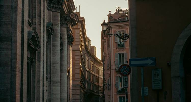 La luz melancólica del atardecer se filtra entre las altas fachadas renacentistas y barrocas de una estrecha calle romana.